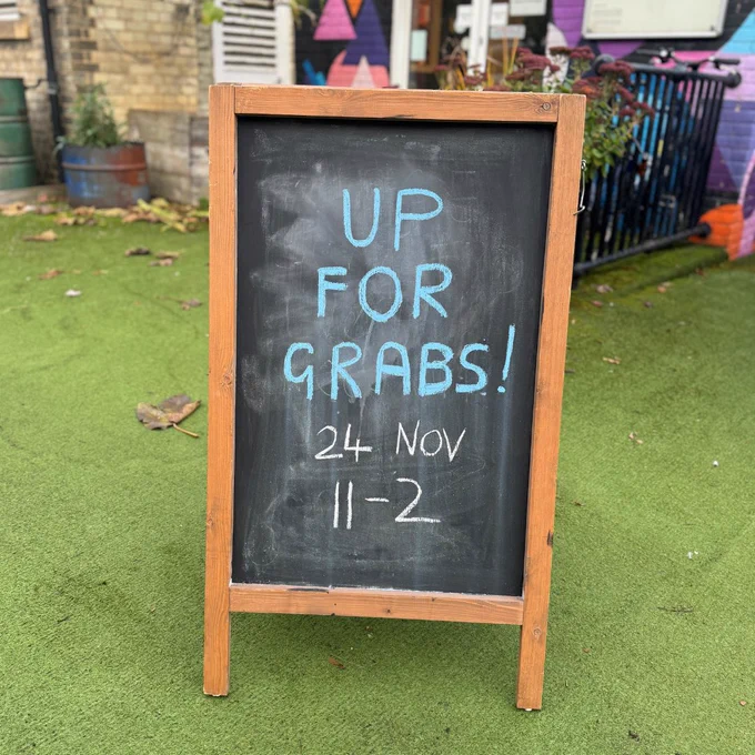 A chalkboard stands in the garden, with blue and yellow chalk text on it which reads ‘Up For Grabs’, ‘24 Nov’, ‘11 -2’.