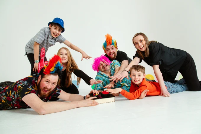 A group of adults and children in colourful wigs and costumes are all reaching for a bright red buzzer on the floor.