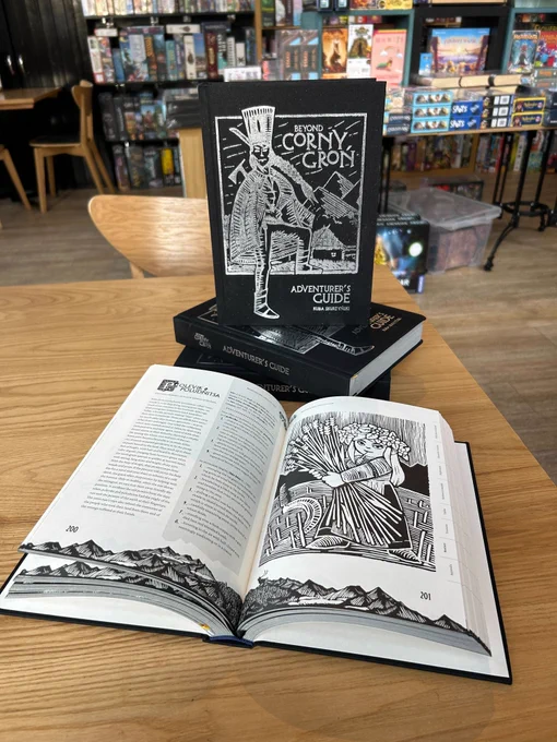 A copy of the RPG Beyond Corny Gron on a table in the Edinburgh board game shop Ancient Robot Games. The book has a dark cover with a woodcarving style illustration of a person standing in front of some mountains.