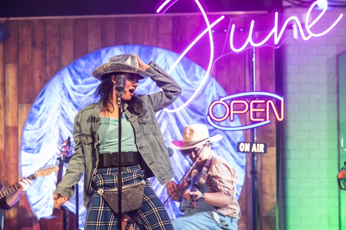 A woman with dark hair is dancing and singing into a microphone, whilst wearing a disco ball cowboy hat with a matching bag. A neon sign in the background reads 'June'.