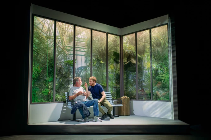 Two men are sitting in front of a glass window packed with plants. One man is pointing in the other mans face. 