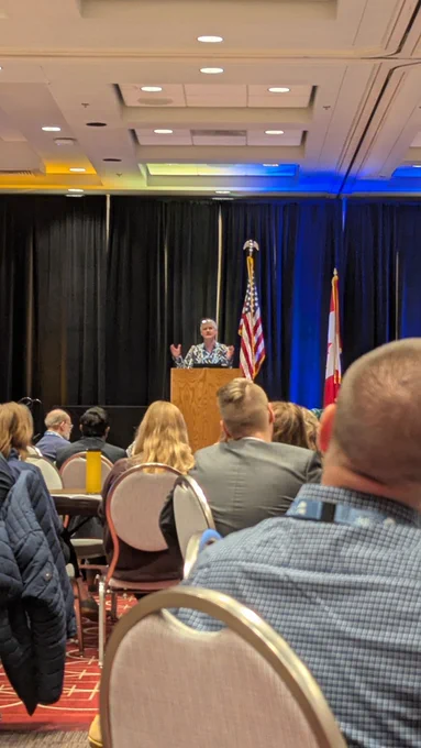 Room of people looking at a man gesticulating at a lectern, wearing a fabulous shirt