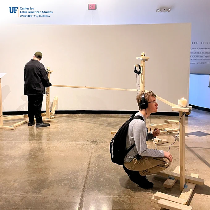 An artwork has three stations with three lit candle and headphones for guests to participate in the artwork. Two attendees are listening - one in the foreground, wearing the headphones, crouched down to look at the candle at eye level.