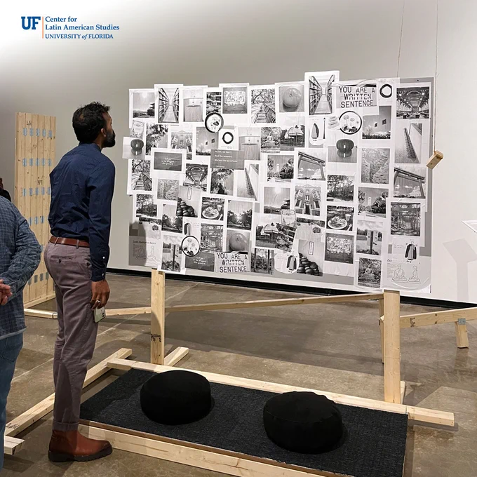 A visitor peers closer at a large installation artwork that has many black and white photos attached to a display board. A wooden block hangs from the ceiling, and two black cushions are presented in front of the display board.