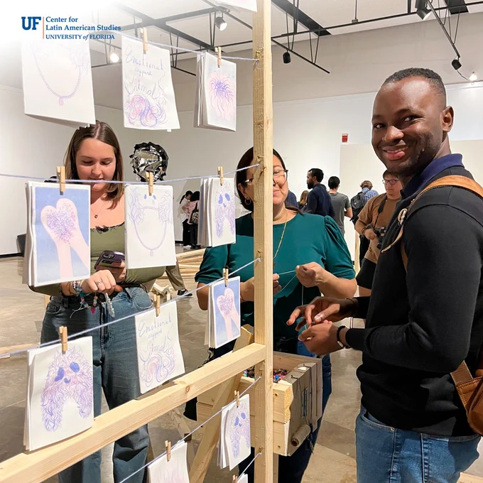 Three students stand in front of an art piece consisting of a wooden frame with rows of string and pieces of illustrated paper clipped to the strong. They are picking out beads to make their own bracelets. One student smiles at the camera.