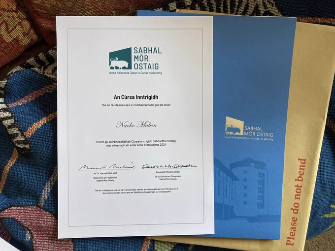 A view of three paper-based items on a colourful soft surface. From the bottom, a light brown envelope, a blue paper holder that has “Sabhal Mòr Ostaig” logo, and a white certificate of a language course. 