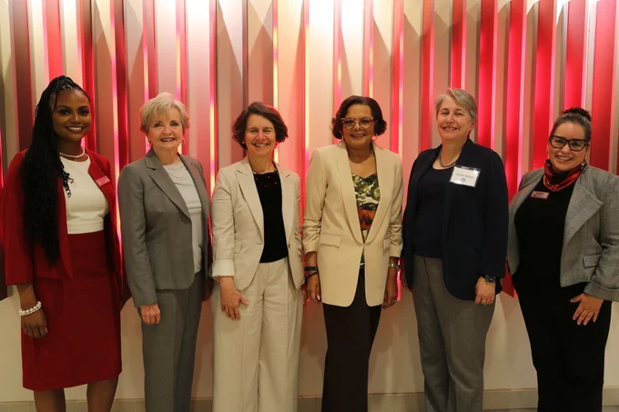 Six women stand in a line in front of a red fin wall. 