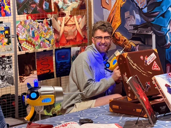 Caleb crouched behind a table and holding a Ripple Gun from Triangle Agency. Around him is a bunch of Triangle Agency art, books, and game boxes. Behind him is a piece of a very large image that hasn't been seen before. Visible is a strange segmented golden limb, a giant blue clawed hand, and a copy machine smaller than the hand. 