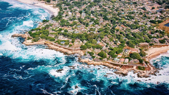 A photo of the California coast featuring a blue ocean and a community of houses and other buildings on land.