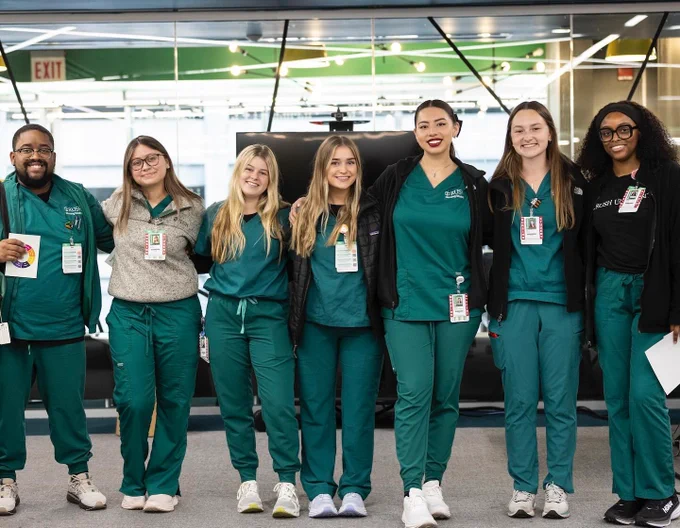 Group of seven Rush nursing students in green scrubs, holding ID badges, smiling and standing together inside a modern building.