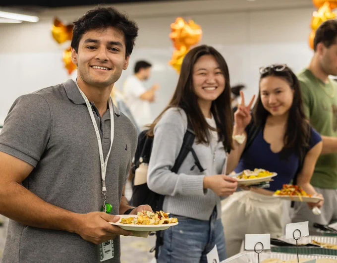 Three Rush University smiling and holding plates of food. One person in the foreground is giving a thumbs up, the second person is smiling broadly, and the third is making a peace sign. They are in a room decorated with gold balloons.