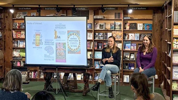 Two people seated in front of a bookshelf in a bookstore, presenting at an event. A projector screen between them displays book covers including titles like "Missing Microbes" and "The Keystone Approach.