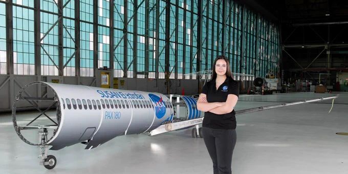 A portrait of Abigail Reigner, standing with her arms crossed in front of a 25% scale model of NASA’s SUbsonic Single Aft eNgine (SUSAN) Electrofan aircraft concept used to test and demonstrate hybrid electric propulsion systems for emission reductions and performance boosts in future commercial aircraft. Credit: NASA/Sara Lowthian-Hanna 
