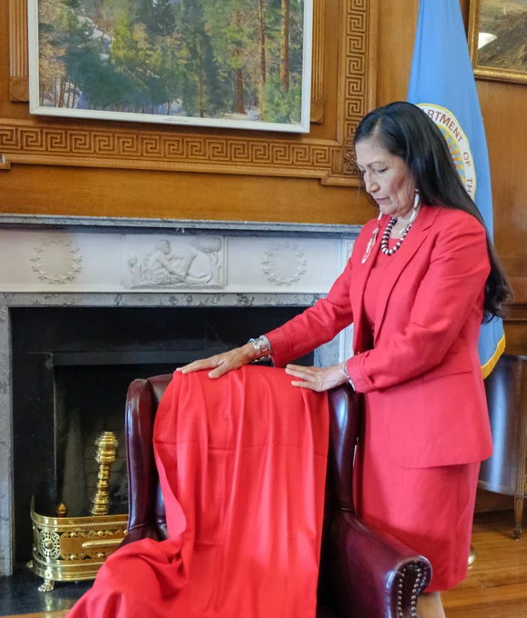 Secretary Haaland wears a red suit and stands next to a leather couch, where she has draped a red shawl. She stands inside a wooden office in front of a fire place.