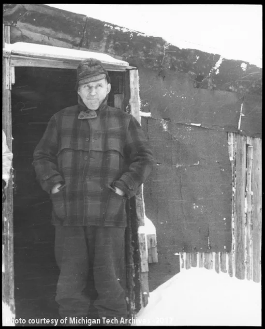 Man in plaid jacket and hat standing in front of a tarpaper house in the snow