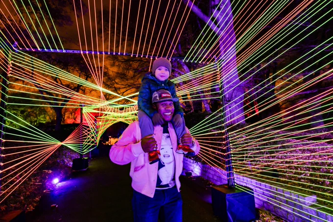 An adult carries a child on shoulders through an art installation made of many glowing strings arranged in a geometric pattern.