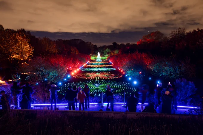 People look from a high vantage point at a long lawn ringed by trees. The lawns has thousands of lights arranged in circular patterns. At the end of the lawn is a pyramid created by lights.