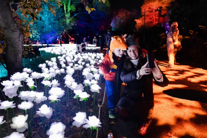 An adult and child in winter wear pose while kneeling on a path in front of a light art installation which consists of hundreds of illuminated white roses.