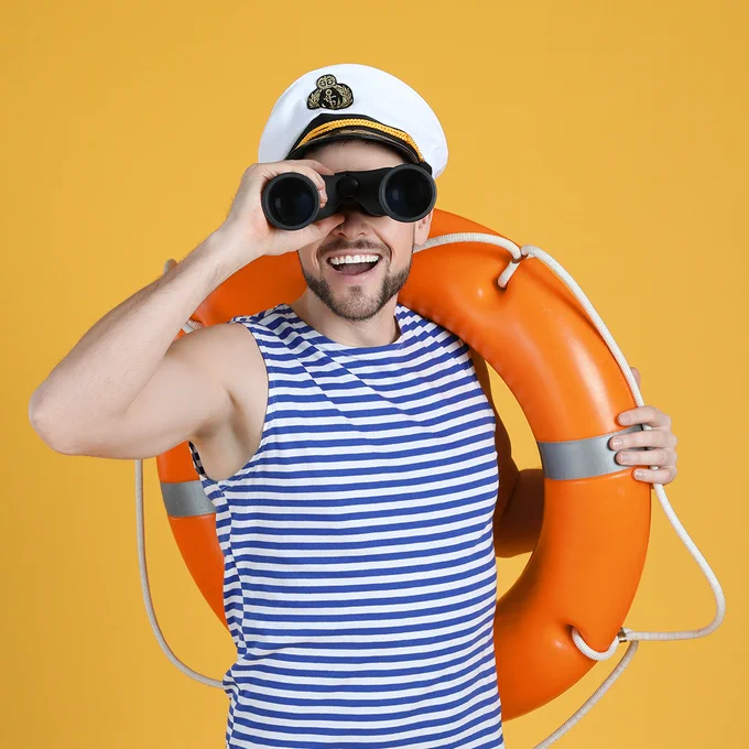 Image of a man in a blue striped sleeveless top, wearing a sailor hat and holding an orange round life preserver, looking through binoculars