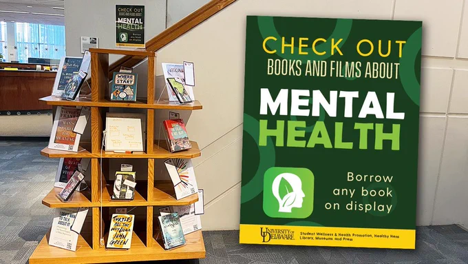 A sign that reads: "Check out books and films about Mental Health" sits in the foreground. A picture of a book display is in the background.