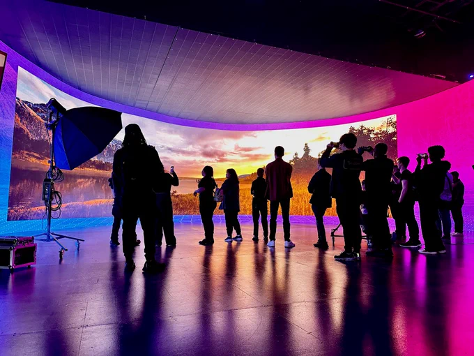 A group of UF Digital Worlds students are photographed standing on a virtual production stage at Vū Technologies in Tampa, Florida. The students are silhouetted against the backdrop of a curved LED volume wall that displays a high-definition, 3D, panoramic view of a lakeside sunrise with a snow-dusted mountain range in the background. Photo by Justin Marlin.