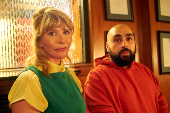 Julia Davis and Asim Chaudhry seated side by side in a warmly lit room with stained glass and framed documents on the wall behind them.