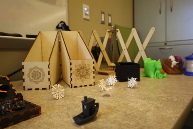 📸: Community Advocate, "Shrewsbury Public Library Unveils Makerspace" by Evan Walsh.  Photo of a desktop with wooden laser cut file folder boxes, 3D printed snowflakes and figurines.