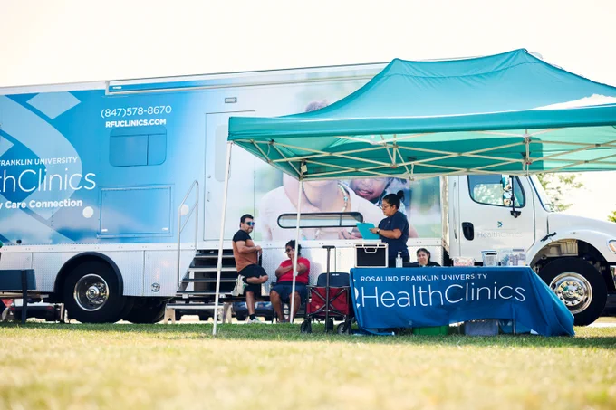 A full view of the Community Care Connection vehicle parked behind a informational table that is set up outside. RFU members sit around the table. A cloth covers the table and reads "Rosalind Franklin University HealthClinics"