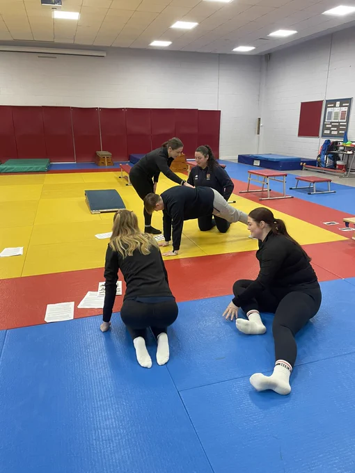 Four trainee PE teachers discussing & practicing gymnastics shapes with Catherine from Positive PE. They’re in the Dojo at Bishop Challoner school with lots of great gymnastics equipment.