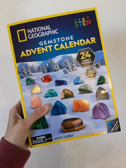 My hand holding the National Geographic GemStone Advent Calendar box. The front of the box has pictures of different gemstones on it.