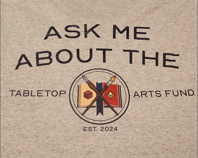 A close-up photo of a t-shirt. The shirt is light grey with black text that reads "ASK ME ABOUT THE TABLETOP ARTS FUND" oriented around a logo. The logo features an ink pen and a paintbrush crossed over an open book, in the style of medieval heraldry. The pages of the book are decorated with a six-sided die and a ten-sided die. 