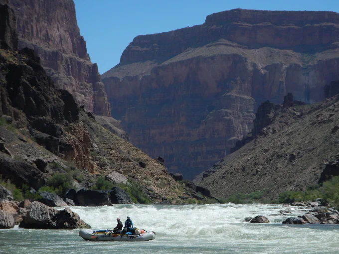A raft travels down the Colorado River with the scenic Grand Canyon in the background.