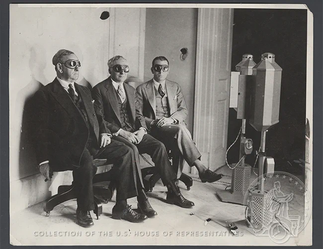 Three men sit in suits and dark goggles in front of old fashioned tanning machines.
