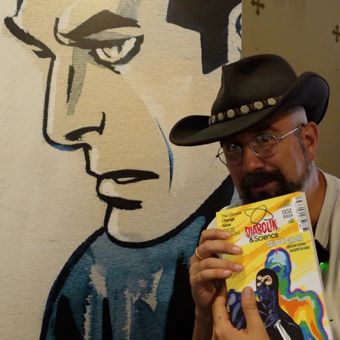 Un uomo sulla destra ha gli occhiali da vista e un cappello da cowboy. Tiene in mano un albo a fumetti intitolato Diabolik&Science e contiene una storia chiamata "Colpo di calore". È Giovanni Eccher, che ha scritto la sceneggiatura dell'albo. Alle sue spalle un pannello con il volto scoperto di Diabolik, disegnato da Giuseppe Palumbo.