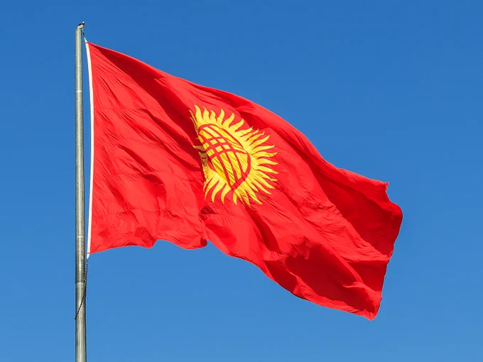 The flag of Kyrgyzstan flying in the wind from the top of a pole on a bright sunny day.