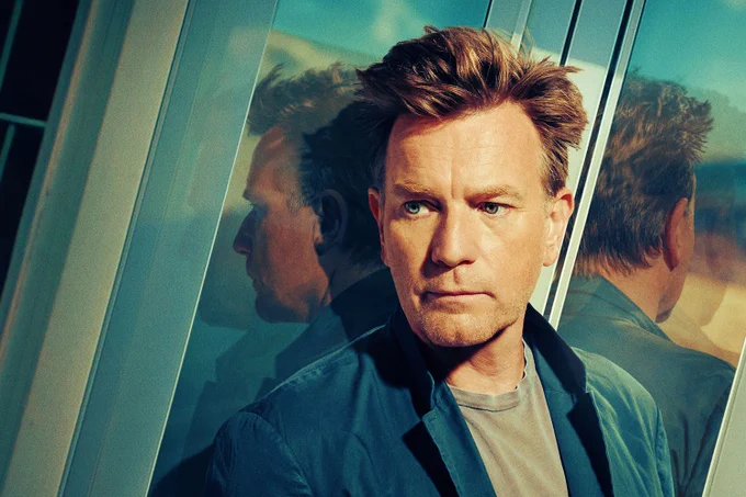 A photo showing a suave man leaning against a french window, wearing a blue shacket and t shirt 