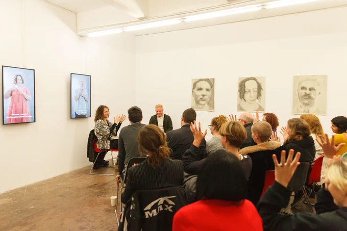 Luke D King and an interpreter sit in front of a crowd of people. There are paintings and digital displays on the walls, which are from Luke's exhibition 'I am a...', 2018. Photo by Snehargo Gnosh