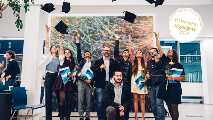 Ein Gruppenfoto. Sieben Personen werfen ihre Abschlusskappen in die Luft. In der Mitte steht ein Professor, ein Glas Champagner in der Hand, mit einem Lächeln im Gesicht. Vorne kniet eine Person ohne Abschlusskappen. Im Hintergrund ist ein großes, buntes wissenschaftliches Bild zu sehen.