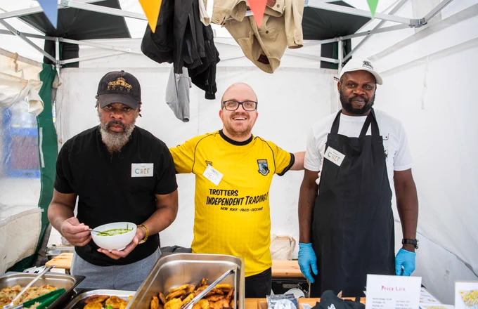 3 men from Headway East London smile for the camera, standing under a gazebo at Hoxton Street Market serving food.