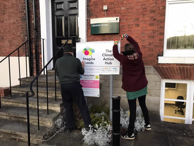 Two people stand outside a red brick building putting up a new white sign which reads ‘Imagine Leeds, The Climate Action Hub’