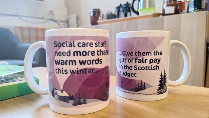 Two mugs sitting side by side on a coffee table. The mugs are illustrated with a winter land and sky-scape scene in purple, green and white. With stars and air currents in the background, the mugs carry a message saying :'Social care staff need more than warm words this winter ... Give them the gift of fair pat in the Scottish Budget.' 