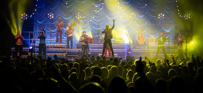 An action shot of Bellowhead live on stage, in the middle of a song, with the audience in the foreground, cast in yellow light, along with more yellow spotlights along the background, in contrast to the blue fabric draped across the back. Of note is John Boden in semi-silhouette who is stood on a platform at the front of the stage, mic to his mouth in song, and left hand raised pointing skyward.