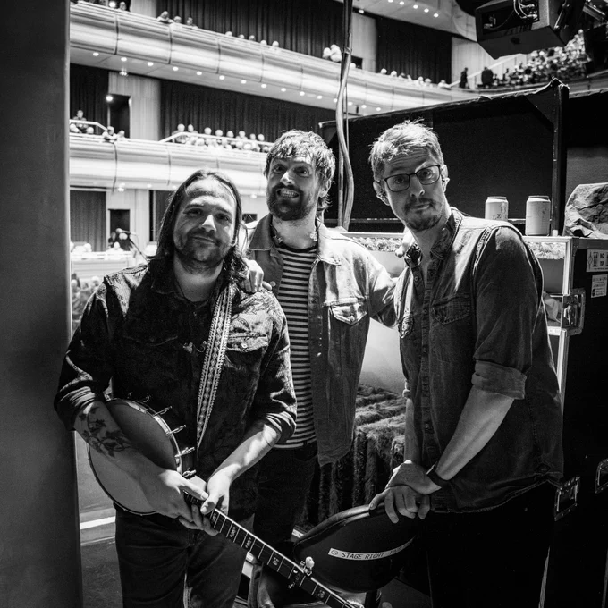 A black and white photo of Andy, Robbie, and JD all in a fairly nonchalant style, looking at the camera. Andy has a banjo slung around his neck, Robbie with a big grin on his face, and JD pulling an "ooh sir" expression whilst leaning on a chair. Behind them is the Dome in Brighton, waiting to be filled later with an audience.