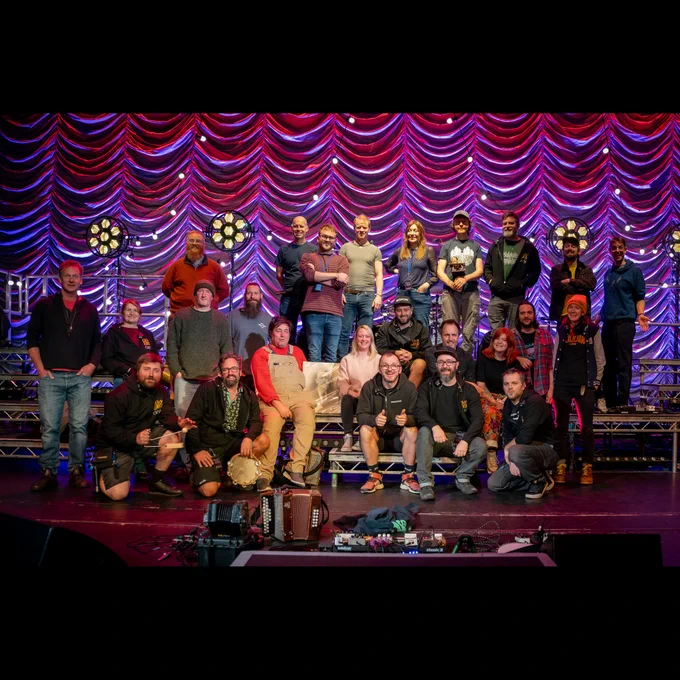 A group photo of both Bellowhead, The Longest Johns, and their various tech crews arranged in three tiers across the stage.
