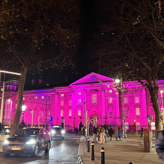 Photo taken at night on College Green with Trinity in the background. The facade of Trinity is lit up in purple for Disability Awareness Week. Cars, trees, and people in the foreground.