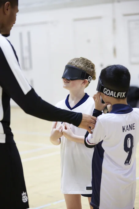 Two blind/vi Bloomsbury Football players wearing England shirts wear blindfolds and listen to coach Owen giving instructions about their next drill. 