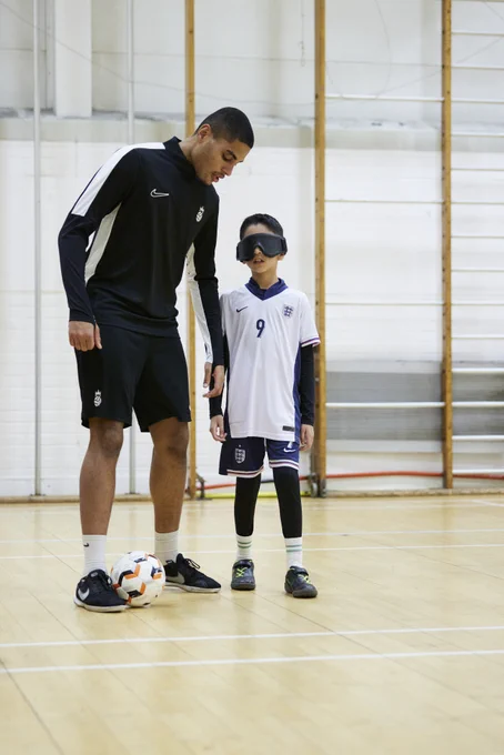 Coach Owen Locke stands next to a blind/vi Bloomsbury Football player. Owen has a football at his feet, and the player is wearing a blindfold. 