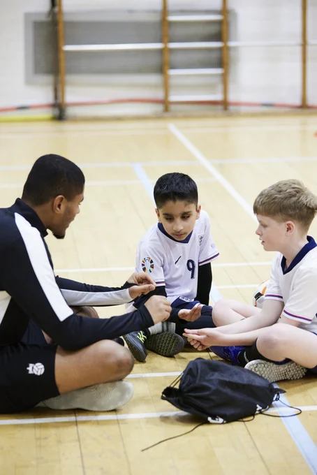 Coach Owen Locke sits on the floor in a circle with two young blind/vi Bloomsbury Football players as they talk football tactics. 