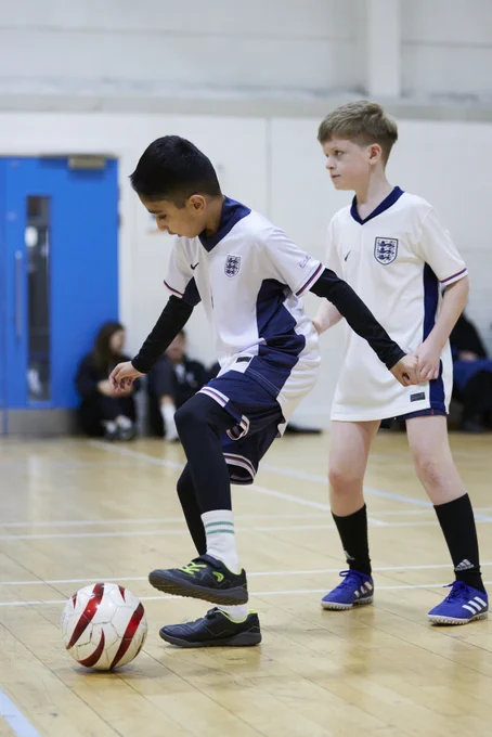 Two blind/vi Bloomsbury Football players are on the pitch playing football. Both are wearing England kit, and one player is about to kick a white and red football. 