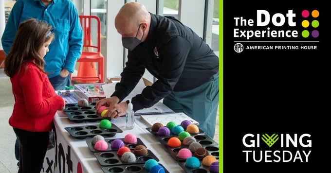 An adult speaks to a young girl from across a table while another man stands at the head of the table. The table between the adults and the child has several muffin tins filled with colorful tennis balls. Text: "Giving Tuesday." The Dot Experience logo.